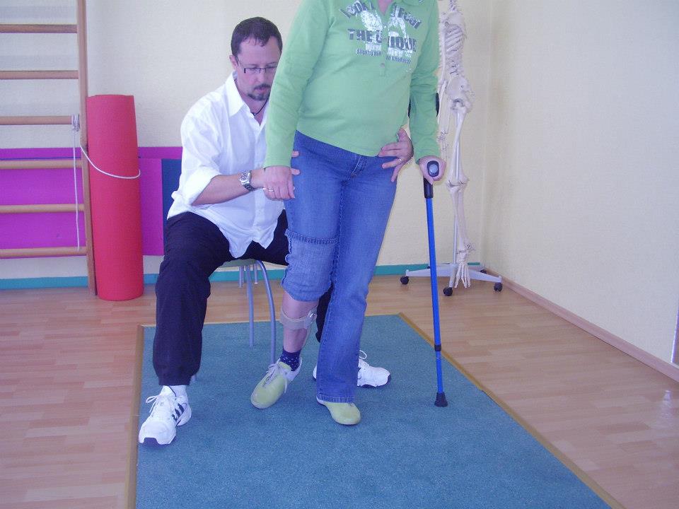 Behandlungen: Das Bild zeigt den Physiotherapeuten Bernd Conrads bei der Behandlung einer Patientin. Er sitzt auf einem Hocker und stützt die Patientin im Hüftbereich. Seine Augen sind auf die Bein- und Fußbewegungen der Patientin sehr konzentriert gerichtet. Die Patientin hält in der linken Hand eine blaue Unterarmgehstütze, auf dem Sie zusammen mit Ihren rechten Bein Ihr Körpergewicht lagert. Ihr rechtes Hosenbein ist bis zum Knie hochgefaltet. Am rechten Unterschenkel befindet sich eine beige Bandage. Der rechte Fuß befindet sich in der Bewegungsphase nach vorn. Sie bewegt sich auf einer blauen langgestreckten Matte. Sie trägt dabei bequeme Straßenschuhe. Im Hintergrund des Bildes steht links eine hölzerne Gymnastikwandleiter, an der eine rötliche Gymnastikmatte aufrecht als Rolle gebunden ist, und rechts ein Skelett auf einem fahrbaren Drehkreuz.